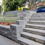 Stone retaining wall with integrated steps in a Sydney garden, surrounded by plants and mulch, supporting a sloped area near a modern house, showcasing expert retaining wall installation services.