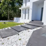 Modern concrete driveway with large, dark paver steps leading to a white house in Sydney, surrounded by white gravel and a well-maintained lawn, showcasing professional concrete driveway solutions.
