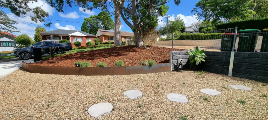 Sydney garden with circular stone pavers set in gravel, featuring a mulched area with plants, a large tree, and metal garden edges, highlighting professional garden edging services.