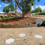 Sydney garden with circular stone pavers set in gravel, featuring a mulched area with plants, a large tree, and metal garden edges, highlighting professional garden edging services.