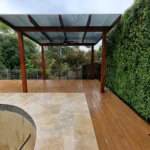 Modern pergola with a wooden deck and tiled area in a Sydney backyard, featuring a green wall backdrop and a metal roof, highlighting professional pergola construction services.