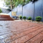 Wet wooden deck with stairs leading to a garden area in Sydney, featuring a dark fence and neatly trimmed bushes, showcasing a professional decking installation service.