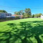 Lush artificial grass lawn in a Sydney backyard, surrounded by trees and a black fence under a clear blue sky, showcasing a professional artificial grass installation service.