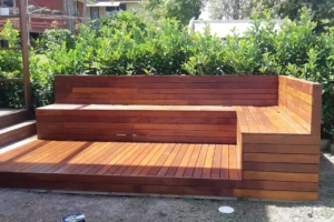 A custom-made wooden garden bench seat by Ariyania Landscapes in Sydney, featuring a sleek L-shaped design on a matching wooden deck, surrounded by lush greenery in an outdoor garden setting.