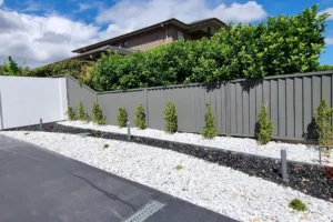 A modern Sydney garden highlights expert garden edging by Ariyania Landscapes. A sleek steel garden edging separates a white pebble pathway from a row of neatly planted shrubs along a gray fence, with black pebbles lining the base of the plants. The setup borders a driveway, with a house and dense greenery in the background under a partly cloudy sky. Explore our metal, steel, timber, corten, plastic, or brick garden edging services for your space!