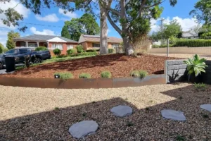 A suburban front yard in Sydney showcases professional garden edging by Ariyania Landscapes. A curved concrete garden edging borders a mulched flower bed with small plants, complemented by a gravel pathway with circular stepping stones. A large tree adds shade, while a driveway with a parked car and neighboring houses are visible in the background under a partly cloudy sky. Contact us for expert metal, steel, timber, corten, plastic, or brick garden edging services!