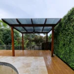 Modern pergola with a wooden deck and tiled area in a Sydney backyard, featuring a green wall backdrop and a metal roof, highlighting professional pergola construction services.
