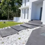 Modern concrete driveway with large, dark paver steps leading to a white house in Sydney, surrounded by white gravel and a well-maintained lawn, showcasing professional concrete driveway solutions.