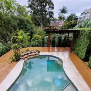Custom timber pool pergola with a transparent roof by Ariyania Landscapes in a Sydney backyard, featuring a wooden deck, tiled pool area, and lush greenery, showcasing swimming pool pergola design.