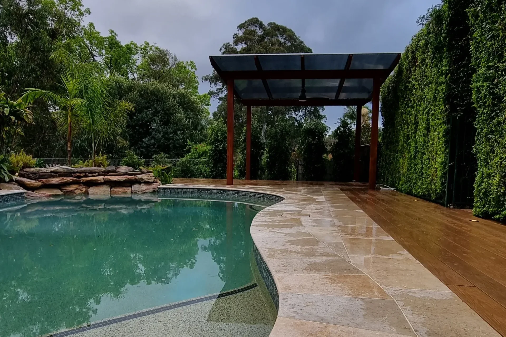 pool landscaping by Arianya, featuring a serene pool with stone paving, a timber pergola, lush greenery, and a rock waterfall, perfect for Sydney backyards.