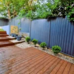 Wet wooden deck with stairs leading to a garden area in Sydney, featuring a dark fence and neatly trimmed bushes, showcasing a professional decking installation service.