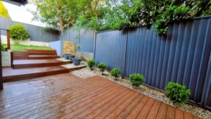 Wet wooden deck with stairs leading to a garden area in Sydney, featuring a dark fence and neatly trimmed bushes, showcasing a professional decking installation service.