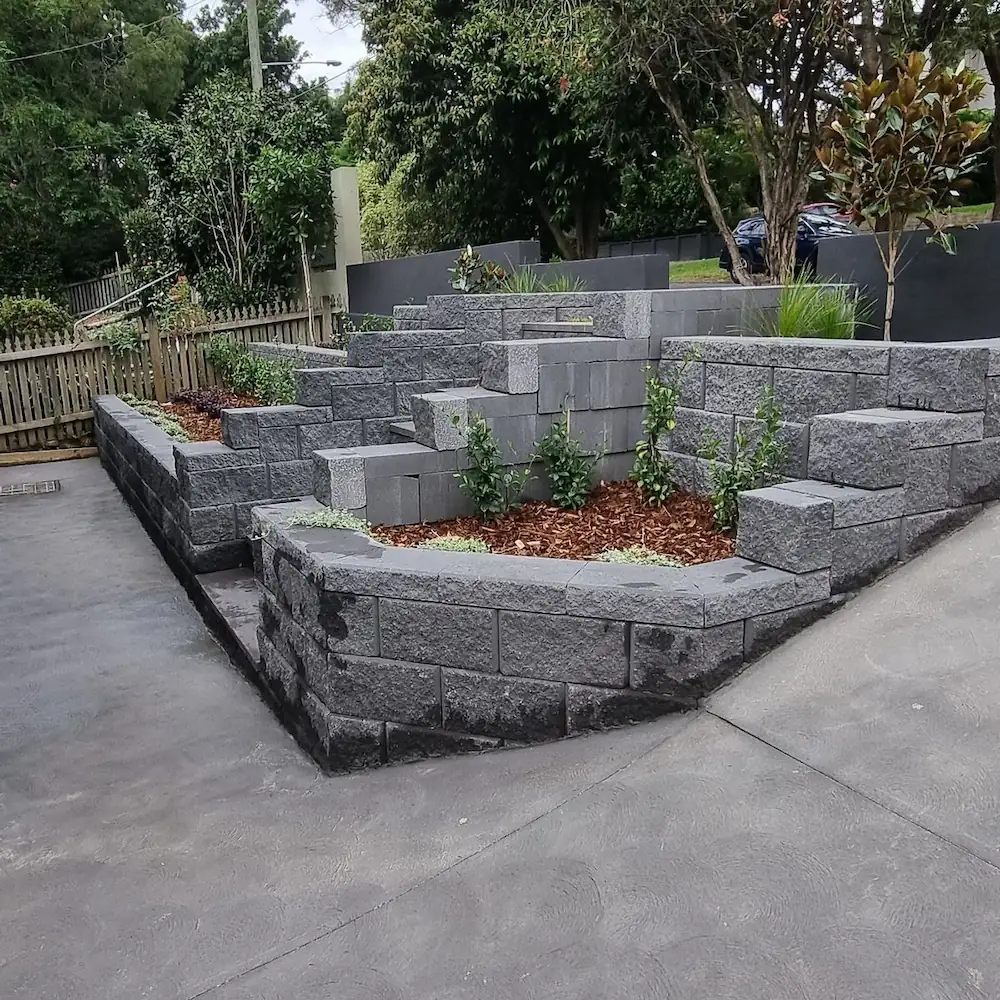 Landscape construction project featuring cement block retaining walls with young shrubs inside and freshly poured concrete ground in a small backyard.